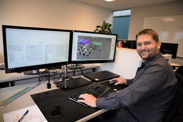 Photo of a smiling professional man sitting at a work station with dual monitors displaying engineering diagrams and 3D modeling software. The desk is equipped with technical drawing tools, a keyboard, a mouse, and laptops, indicating a technical or engineering work environment. The office has a whiteboard with written notes in the background, suggesting an active project or collaborative work.
